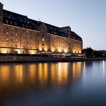 Copenhagen Admiral Hotel Exterior foto The photo shows a large, historic building located beside a calm body of water, likely a river or canal. The structure features a series of tall windows, a dark roof, and is illuminated by warm lights, creating a cozy atmosphere at twilight. Reflecti