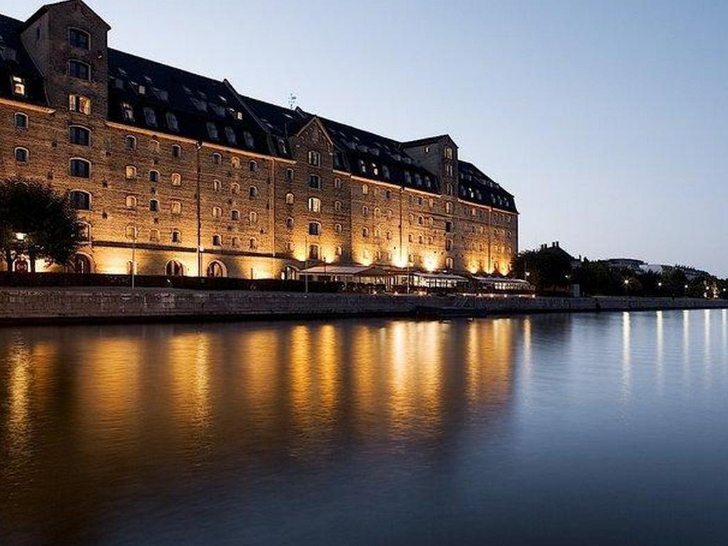 Copenhagen Admiral Hotel Exterior foto The photo shows a large, historic building located beside a calm body of water, likely a river or canal. The structure features a series of tall windows, a dark roof, and is illuminated by warm lights, creating a cozy atmosphere at twilight. Reflecti